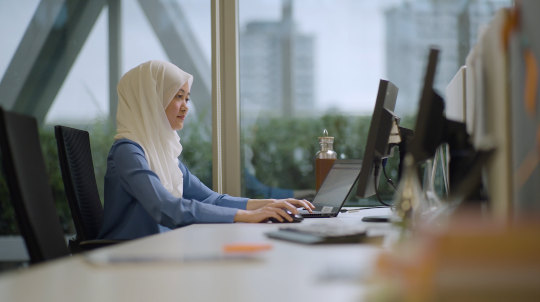 A woman in a white headscarf and blue shirt works on a laptop at a cluttered desk with a window and cityscape in the background.
